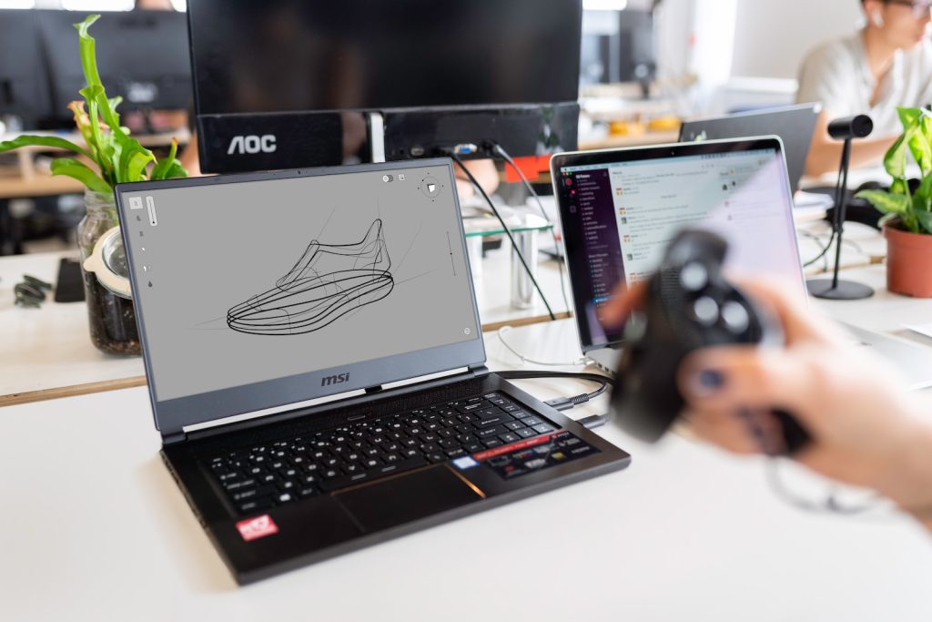 A laptop displaying a 3D shoe design sketch next to a person holding a 3D mouse in a modern office